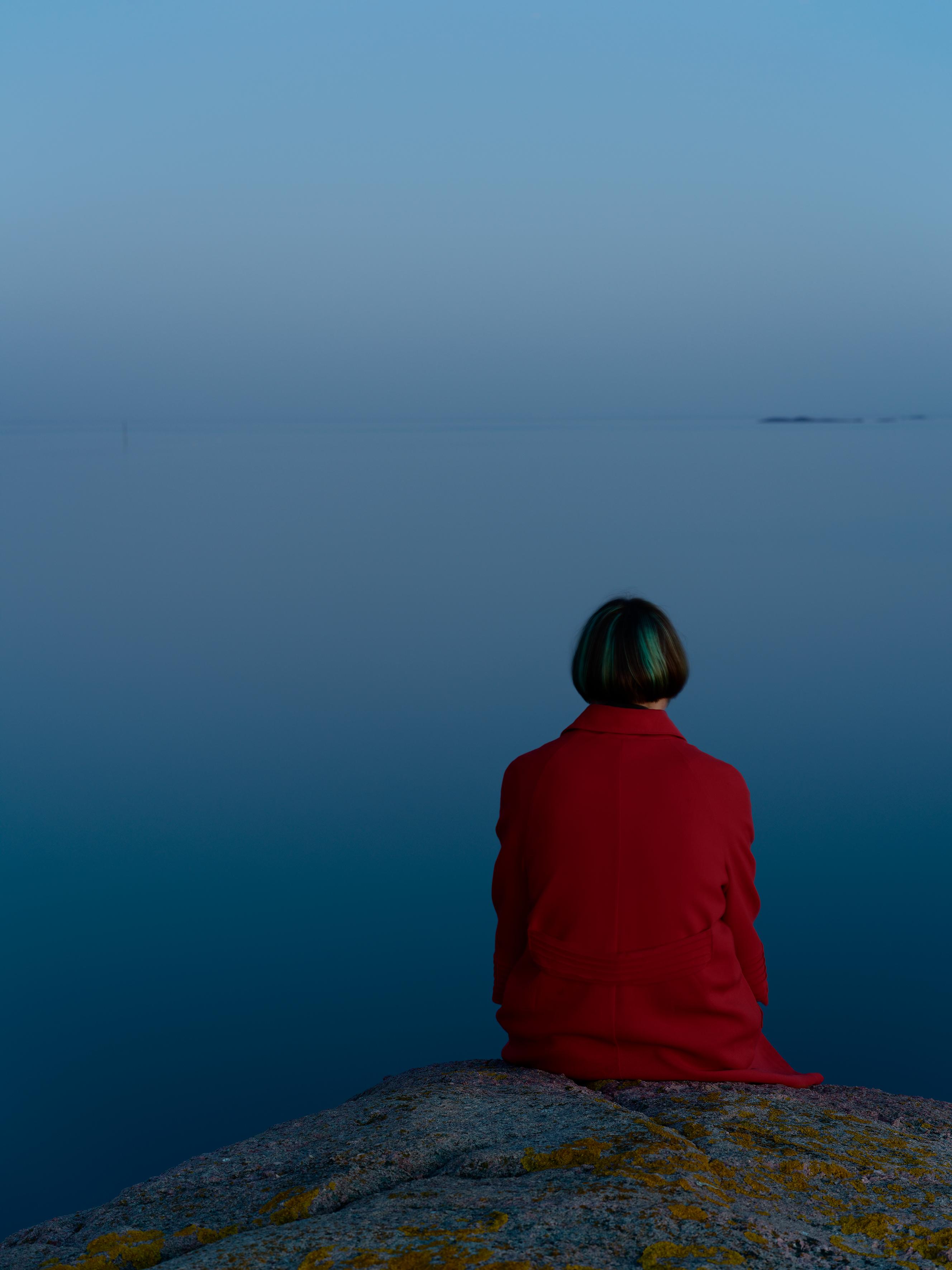 Kvinne i rød kåpe sitter på et svaberg ved havet